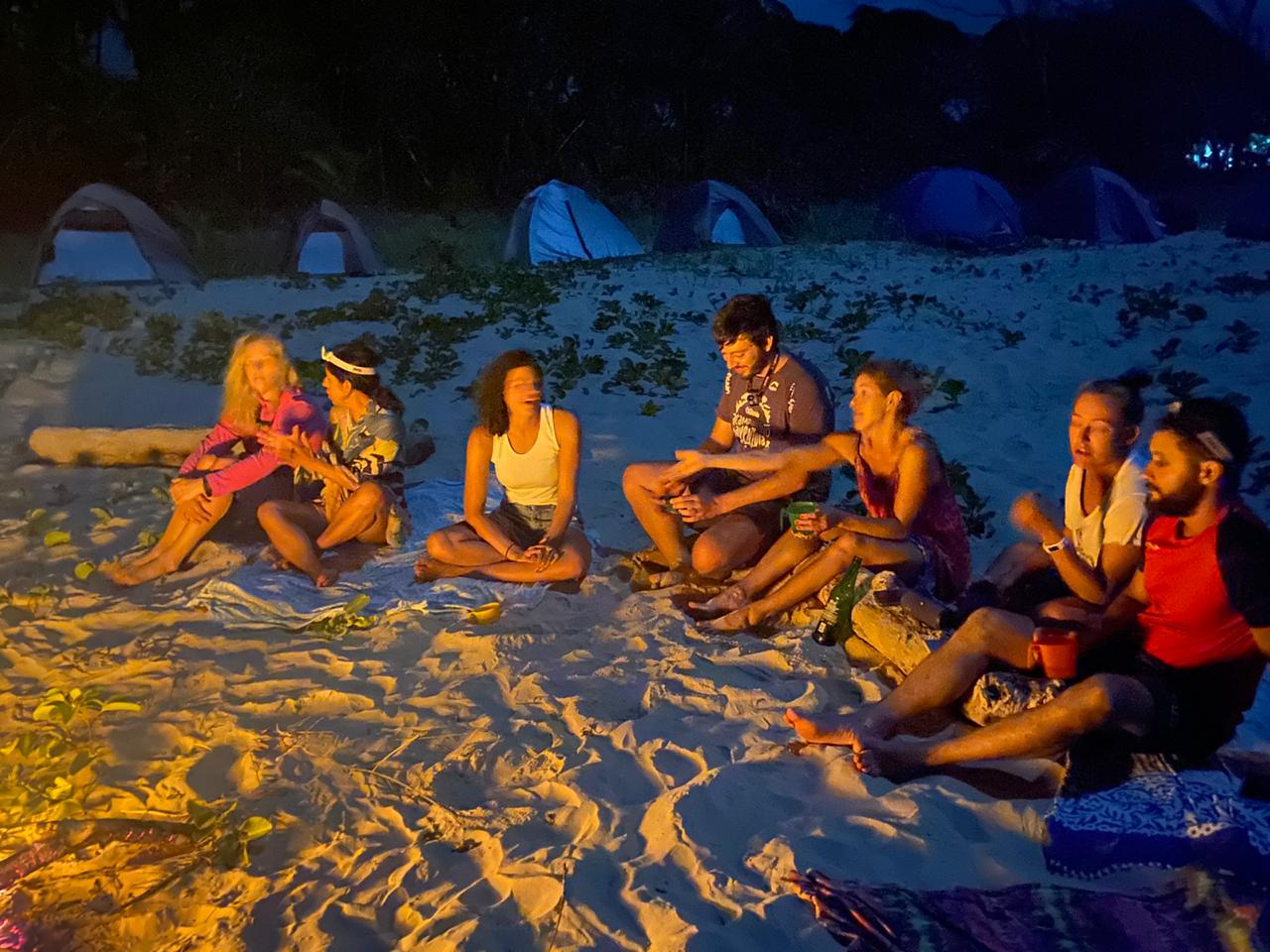 Grupo reunido ao redor da fogueira durante acampamento na praia à noite