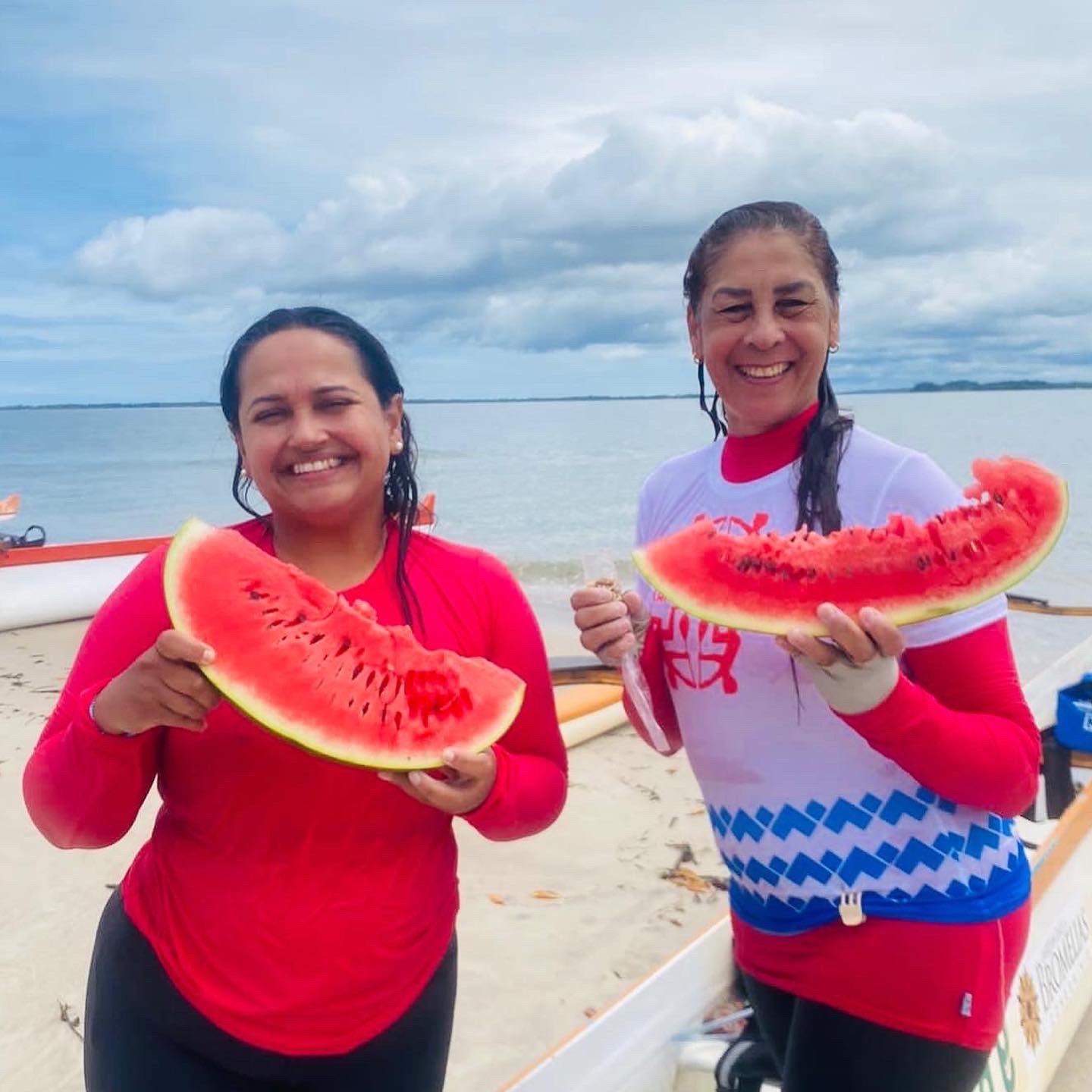 Duas remadoras sorrindo com fatias de melancia na praia